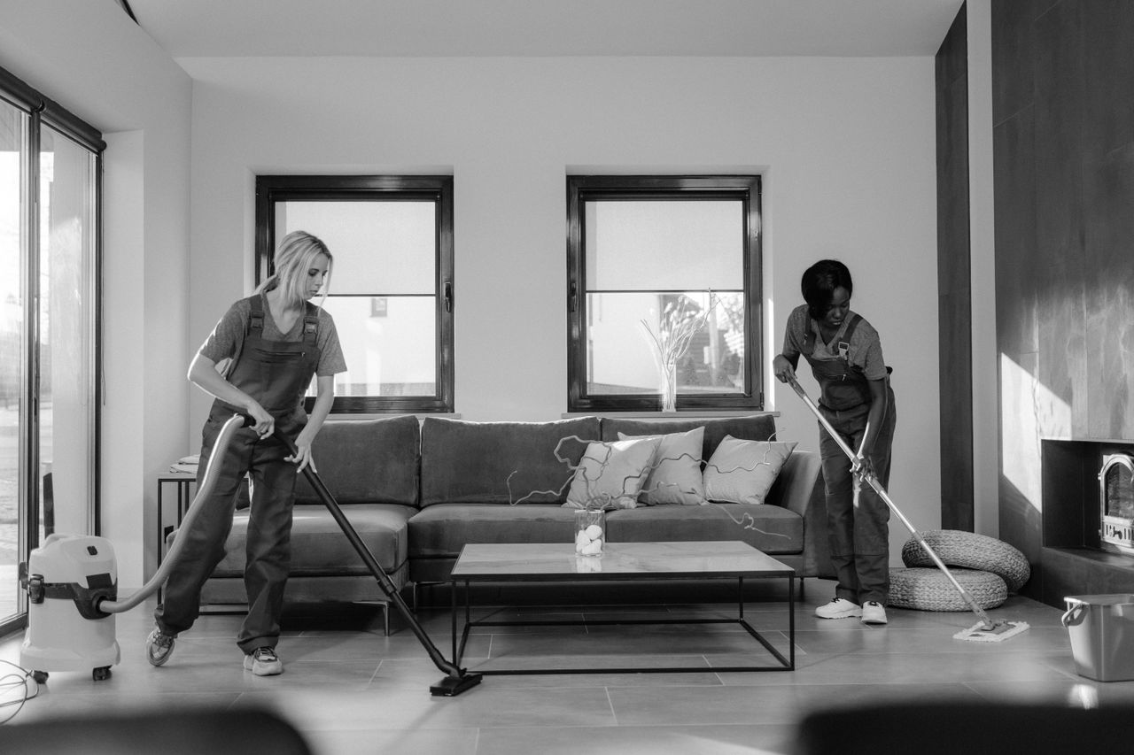 about-img-01 Two women in work clothes cleaning a modern living room using a vacuum and mop.