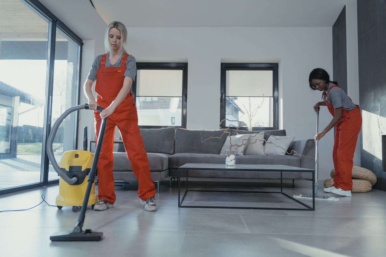 journey-02 Two women in red overalls vacuuming and sweeping a modern living room.